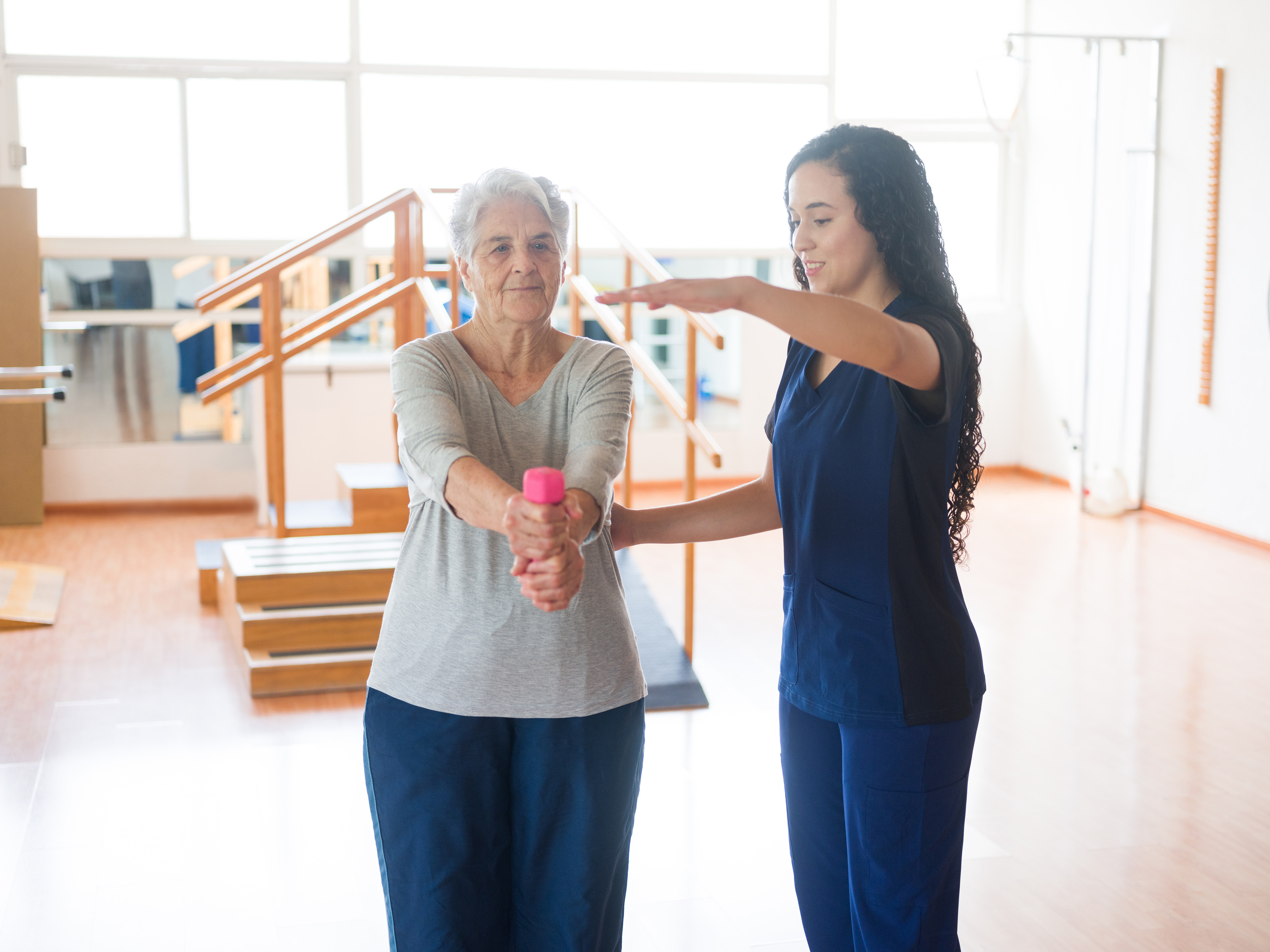 Senior woman lifting weight with both arms next to occupational therapist